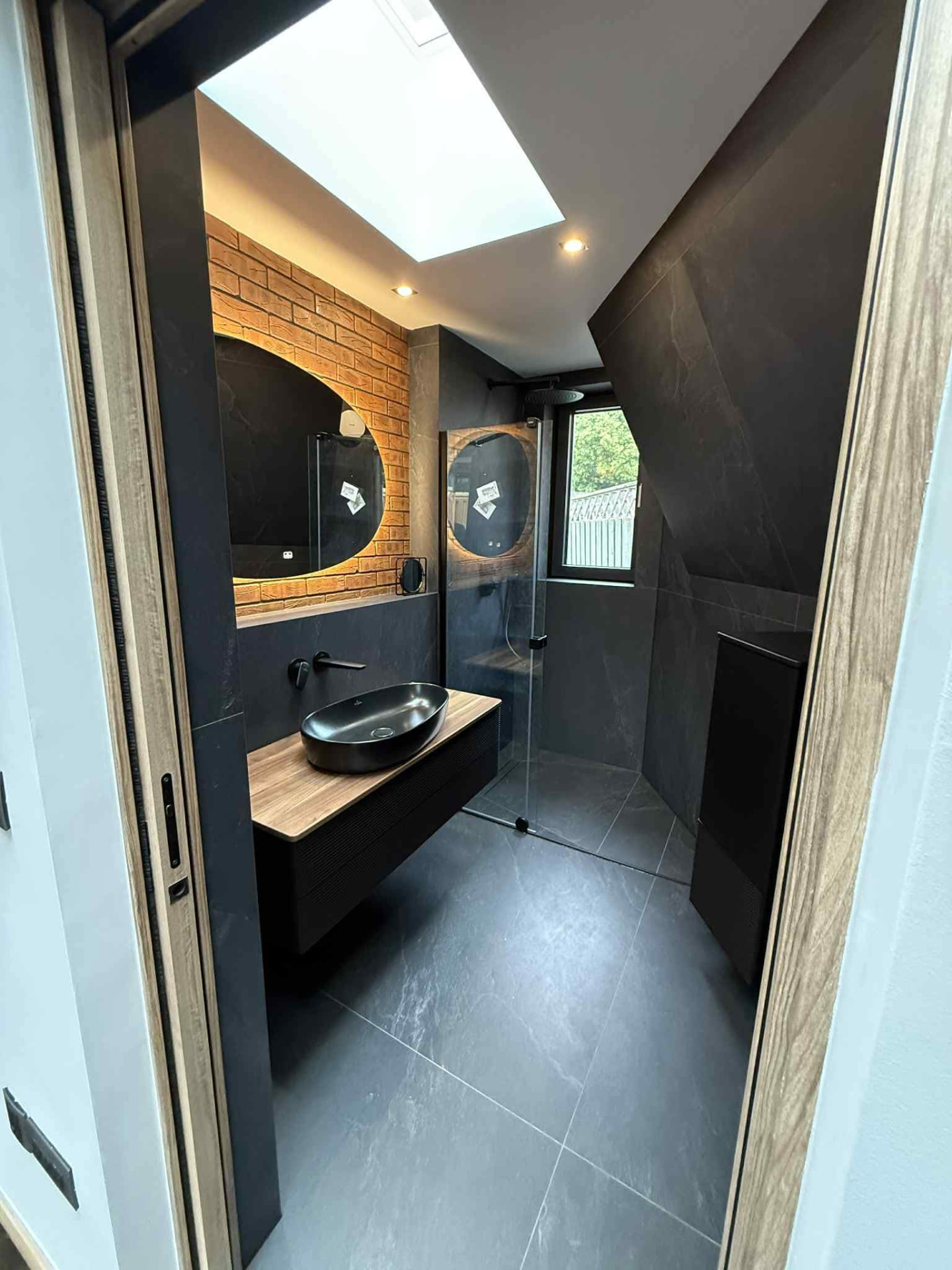 Dark tiled bathroom with skylight, glass shower and round mirrors